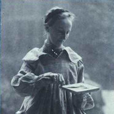 Mary Watts in a long dress and apron, holding a small palette, looking down with a focused expression.