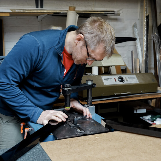 A craftsman meticulously cuts a frame component using specialized equipment in a busy framing workshop, surrounded by materials and samples, demonstrating the precision involved in custom framing.