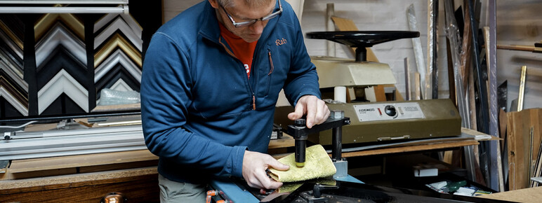 A craftsman meticulously cuts a frame component using specialized equipment in a busy framing workshop, surrounded by materials and samples, demonstrating the precision involved in custom framing.