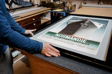 A man framing a poster of a ship, titled "Normandie"