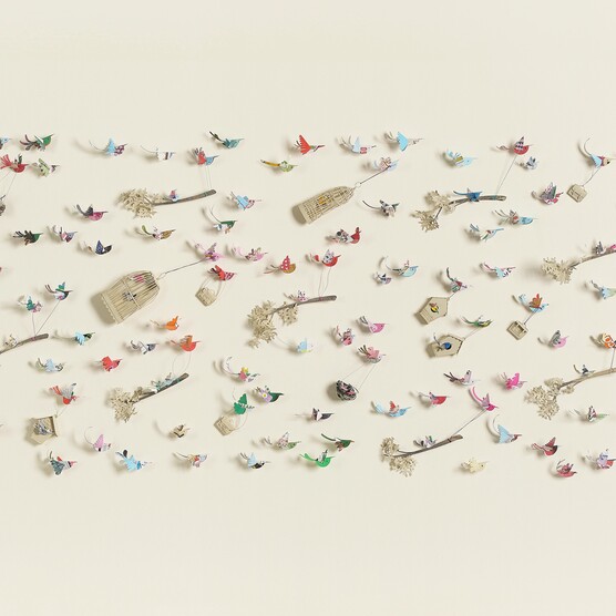 A flat, rectangular artwork against a light beige background, featuring numerous small, colourful paper birds in flight. Some birds are perched on thin branches with delicate white clusters, while others are suspended mid-air with wire-like attachments. Scattered amongst the birds are small birdhouses and cages. The title, "Crowded Skies," by Lucy Dorothy