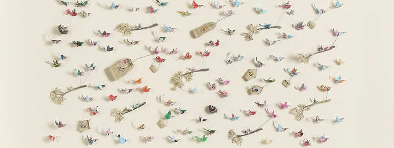 A flat, rectangular artwork against a light beige background, featuring numerous small, colourful paper birds in flight. Some birds are perched on thin branches with delicate white clusters, while others are suspended mid-air with wire-like attachments. Scattered amongst the birds are small birdhouses and cages. The title, "Crowded Skies," by Lucy Dorothy