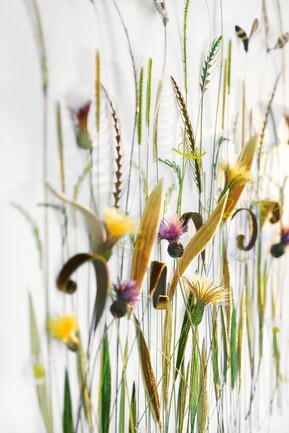 Close-up shot of a stylized floral display against a white background. The arrangement includes various grasses, stylized yellow and purple flowers, and decorative elements like curved brown velvet-like shapes and thin green and yellow wire. The composition evokes a natural yet artistic feel, with soft focus and varying depths creating an ethereal effect.