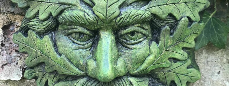 A green, weathered stone sculpture of a Green Man with a face framed by oak leaves, eyes are looking forward, framed by leaves, and partially covered with live ivy, mounted on a weathered brick wall.