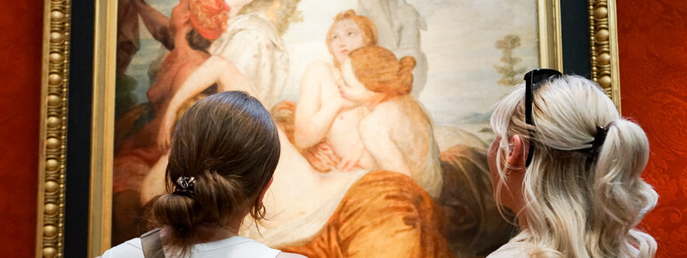 Close-up photo of two women looking at a classical painting. The woman on the left had dark hair in a bun and the woman of the right has blonde curly hair with sunglasses on her head.