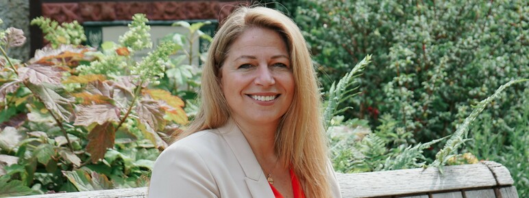 Photo of a woman with medium-length blonde hair wearing a cream-coloured blazer and coral coloured blouse. She is sitting on a wooden bench and smiling at the camera.