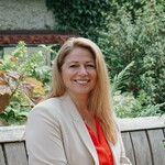 Photo of a woman with medium-length blonde hair wearing a cream-coloured blazer and coral coloured blouse. She is sitting on a wooden bench and smiling at the camera.