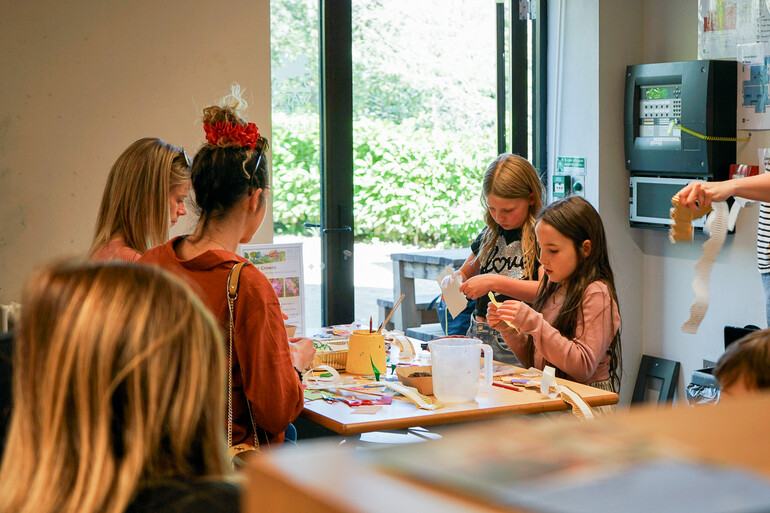 Photo of two young girls making paper crafts at a table in a workshop as their mothers sit facing them.
