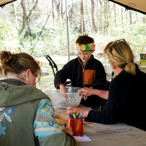 Photo of a young boy under a tent wearing a paper crown, mixing soil in a bowl with a woman either side of him.