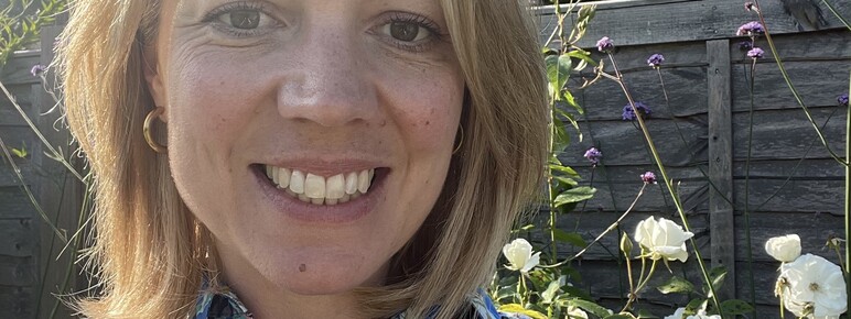 A smiling woman with blonde hair and freckles wearing a vibrant blue floral shirt, taking a selfie outdoors in a sunny garden.
