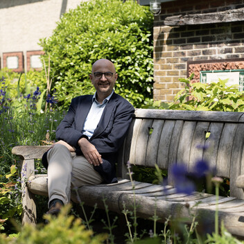 A middle-aged man with a bald head and glasses, dressed in a blue collared shirt, dark blue blazer, and light beige trousers, sits comfortably on a weathered wooden park bench. He is smiling and looking directly at the camera. He is surrounded by a lush green garden with purple flowers in the foreground and background, with a brick wall and green foliage behind him under bright sunlight.
