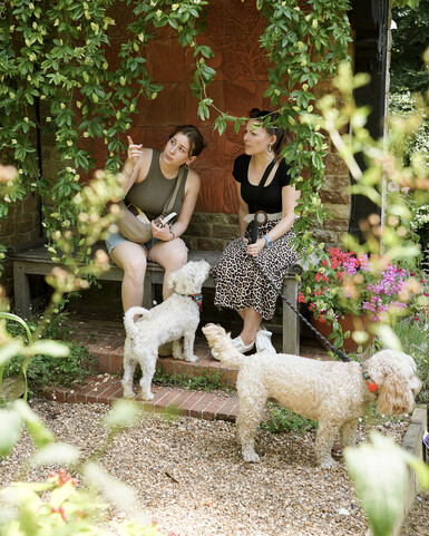 Two women are seated on a rustic wooden bench under a brick archway draped with flowering green vines. The woman on the left, with dark hair in a bun and wearing a green top and shorts, points upwards to the left. The woman on the right, also with dark hair and wearing a black top and a leopard-print skirt, holds a dog leash. Two small, light-coloured fluffy dogs stand on a gravel and brick path in front of them. Red and pink flowers are visible to the right.