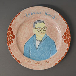 A ceramic plate decorated with a painting of Lubaina Himid. The plate is resting on a grey background. The plate is pale orange with circles of darker orange around the edge. The portrait of Himid sits in the centre of the plate, depicting a women looking off to the side, she is wearing a light blue long sleeve top and orange glasses. Above the portrait it reads 'Lubaina Himid' and below at the bottom of the plate is says 'We will be'.