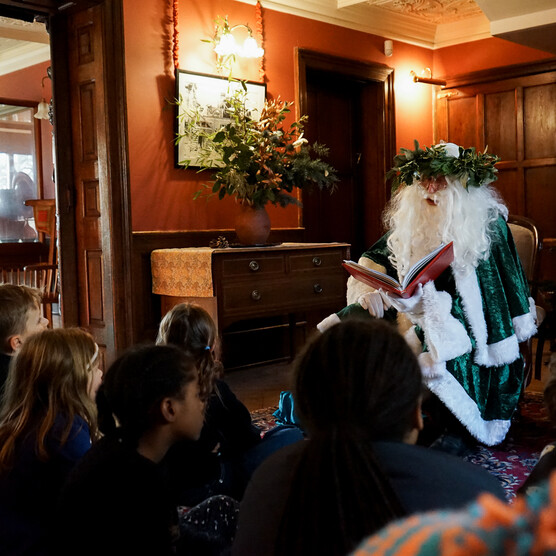 Photograph inside Limnerslease of Father Christmas dressed in traditional Victorian green, reading a story to a group of children sat down in front of him. There is a decorated Christmas tree to the right of Father Christmas