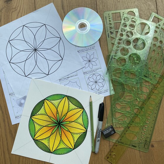 A table with a green and yellow draw mandala. Surrounded by drawing tools, a pencil, pen, sharpener, stencils and a disc.