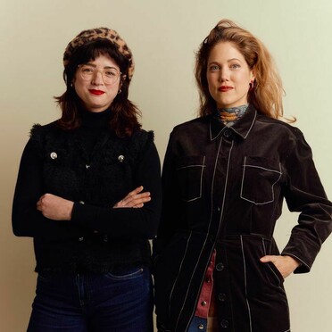 Two women posing together; one wears a leopard print beret, glasses, black turtleneck, a black vest with silver buttons, and dark blue jeans; the other wears a long brown coat with white stitching, a patterned scarf, and red earrings.