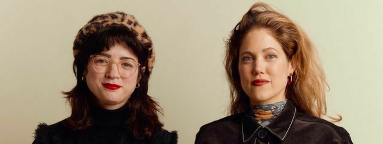 Two women posing together; one wears a leopard print beret, glasses, black turtleneck, a black vest with silver buttons, and dark blue jeans; the other wears a long brown coat with white stitching, a patterned scarf, and red earrings.