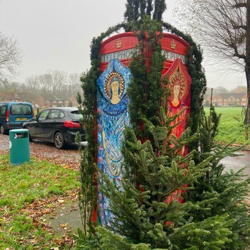 A red phone box covered by life size images of angels from Watts Chapel, decorated with collage and surrounded by fir branches