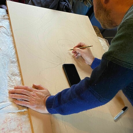 A man leans over a piece of plywood, drawing the outline of an angel from a photo taken in the Watts Chapel