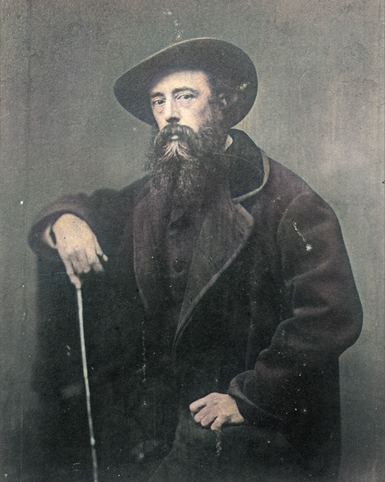 A colour photograph of George Watts posing for his photo wearing a large coat, a hat and holding a stick