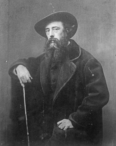A black and white photograph of George Watts posing for his photo wearing a large coat, a hat and holding a stick.