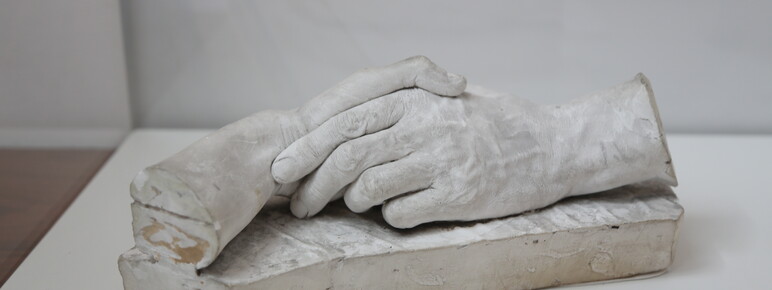 A plaster cast sculpture of intertwined human hands resting on a rectangular base, displayed in a museum setting with an informational label visible below.