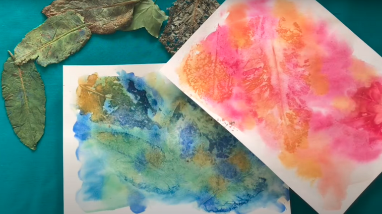 Two watercolour paintings with abstract patterns, one with warm red and orange tones, and another with cool blue and green shades, surrounded by assorted leaves on a light surface.