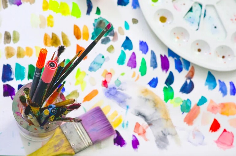 A vibrant image of an artist's workstation with paintbrushes in a jar, a palette with various colours, and scattered colourful paint swatches across a white surface.