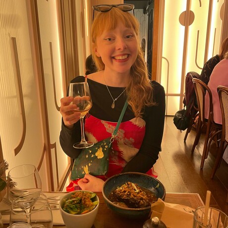 A woman with long red hair sits at a small table in a Paris cafe with a glass of wine and a meal