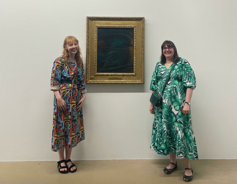 Two women stand either side of a framed oil painting on a white wall