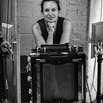 Photo of a woman smiling at the camera with a large format camera in the foreground in front of her.