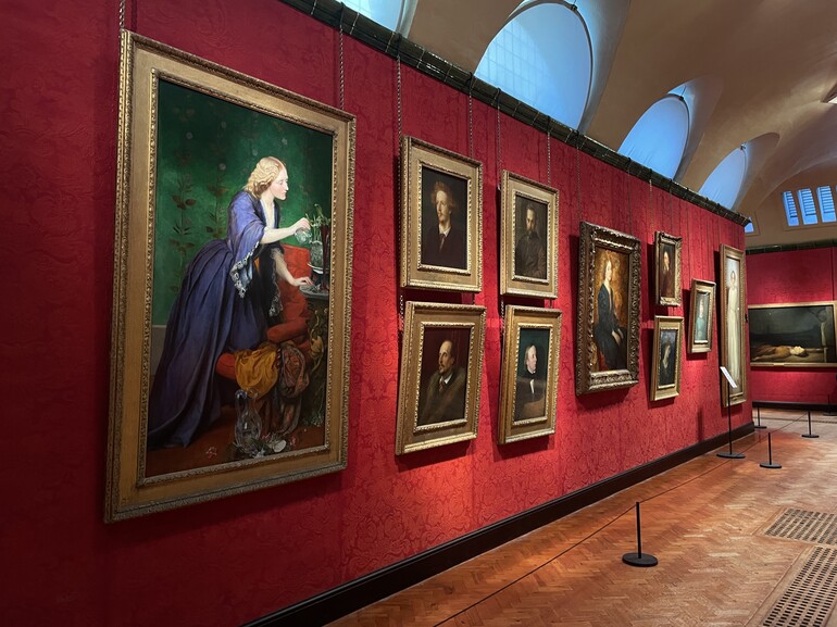 A full-length portrait of female, turned to right, in an informal blue bell-shaped dress with no undersleeves and low-cut neck, kneeling on crimson armchair, watering lilies of the valley on gilt console table. Flowers and carafe in foreground to right; in the background green wallpaper with floral pattern. This painting is placed within the Historic Galleries at Watts Gallery, which has red wallpapered walls and features portraits of men beside her.