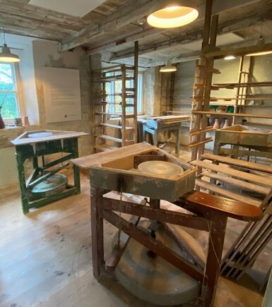 Photo of a pottery studio with wooden kickwheels placed in the middle of the room.