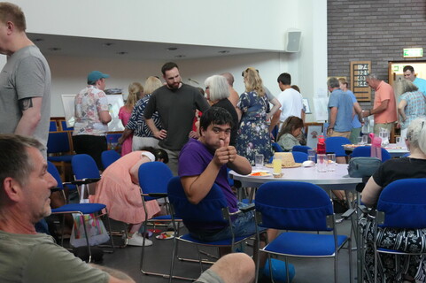 people are lining up at the edge of a busy room, several people are sitting around circular tables in the centre and one man in a purple t-shirt is looking at the camera giving a thumbs up