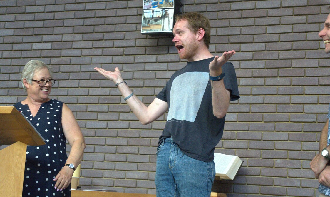 A man in a black and white t-shirt is holding is arms up and smiling broadly. A lady in a blue polka dot dress is looking at him and smiling