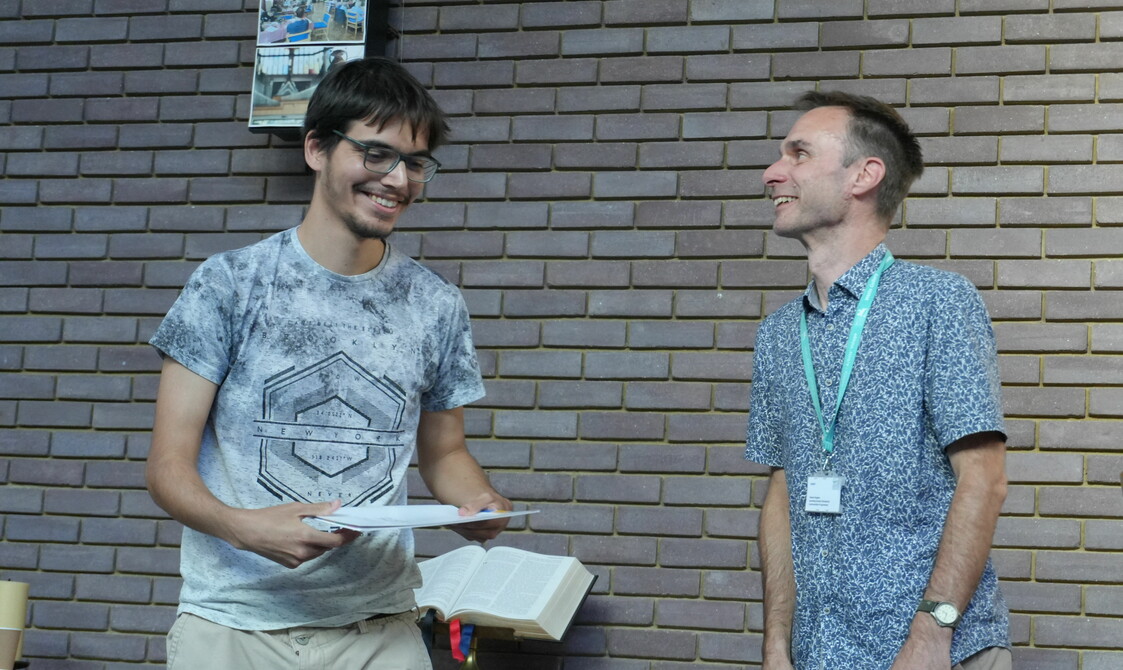 A man in a t-shirt and glasses is smiling broadly as he holds a certificate another man in a shirt is smiling