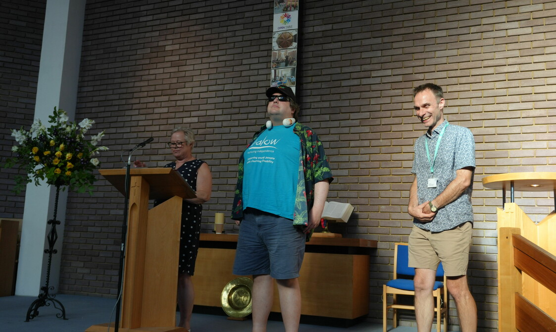 A man in a blue t-shirt and colourful shirt stands on a stage next to him is a woman at a lectern and a man who is smiling