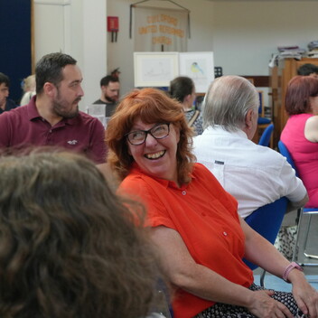 A woman in a red top and black glasses is looking over her shoulder and smiling. She is sitting in a room full of people