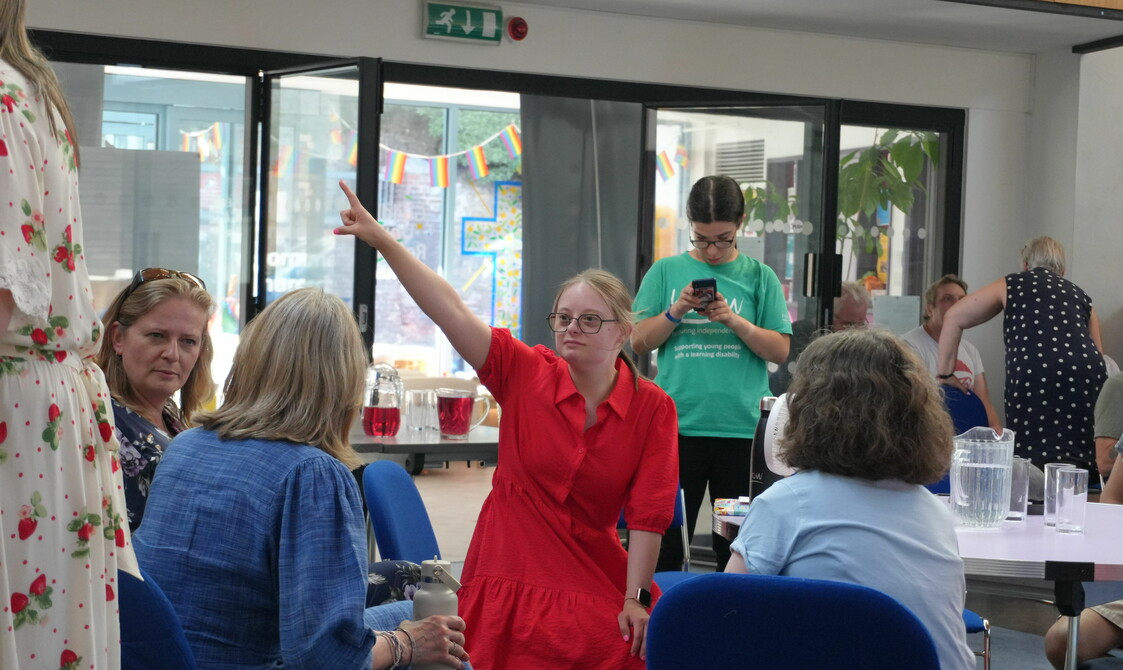 A group of women are sitting on chairs facing each other and talking. A lady in the centre in a red dress is pointing up to the corner.