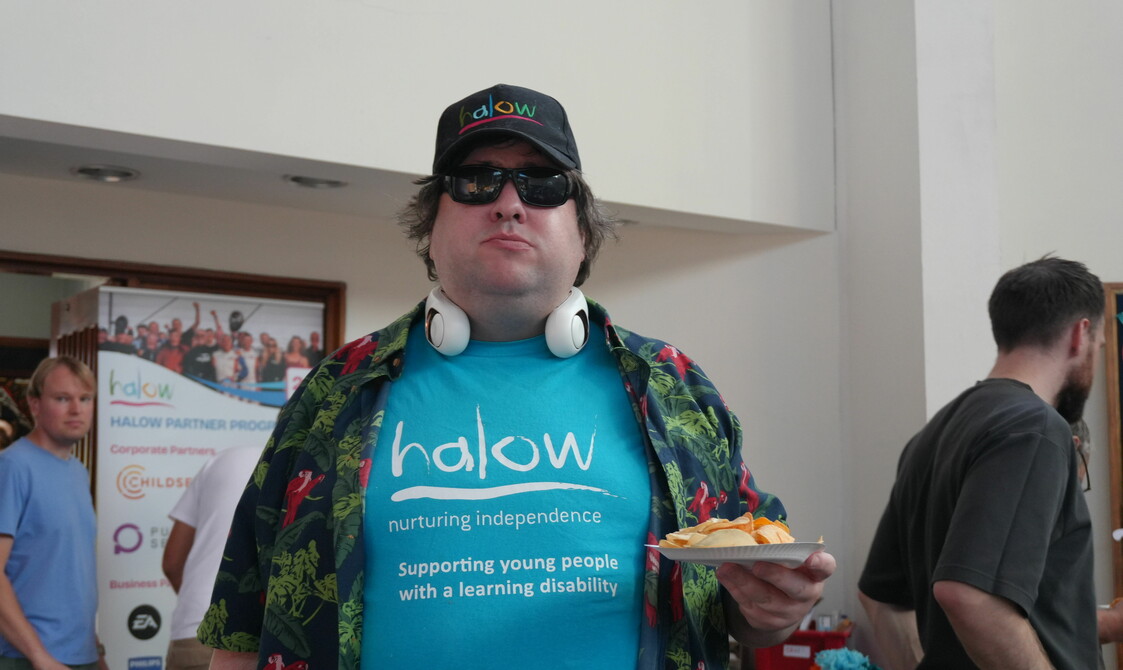 A man faces the camera, he is wearing a black baseball cap and black sunglasses. His blue t-shirt has the word 'halow' written across the front and he has a green and red shirt over the top. He is holding a small paper plate of crisps