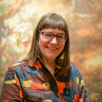 Laura wears a colourful shirt and stands in front of a painting