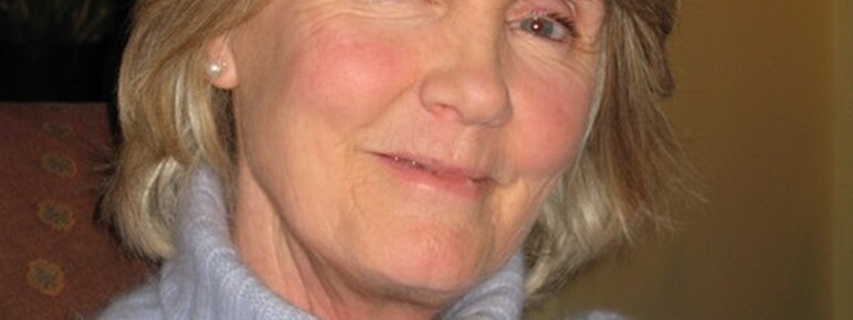 A photograph of a woman looking at the camera smiling, with short blonde hair wearing a blue jumper