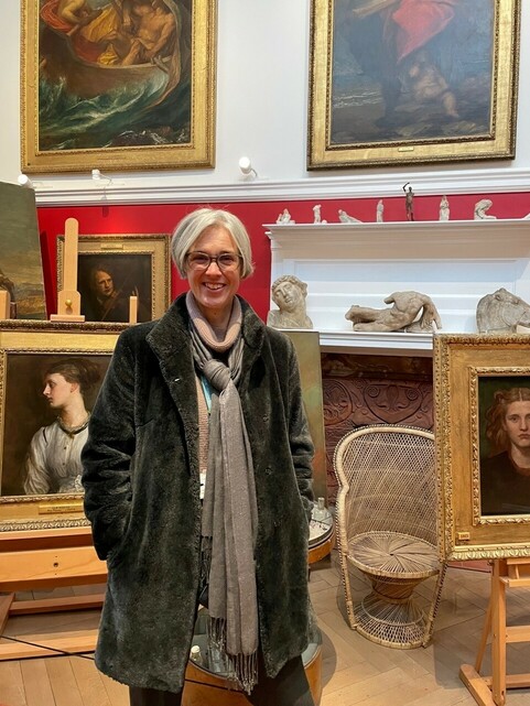 A photograph of Celia stood in George Watts's studio looking at the camera smiling with short grey hair, wearing glasses