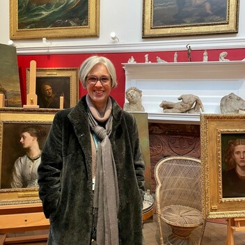 A photograph of Celia stood in George Watts's studio looking at the camera smiling with short grey hair, wearing glasses