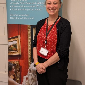 A woman is standing in front of a Watts Gallery roller banner. She is wearing a red shirt and black trousers and is smiling