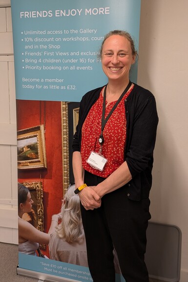 A woman is standing in front of a Watts Gallery roller banner. She is wearing a red shirt and black trousers and is smiling