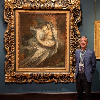 A wearing glasses and a blue jacket is standing next to a large painting by George Watts of Paolo and Francesca