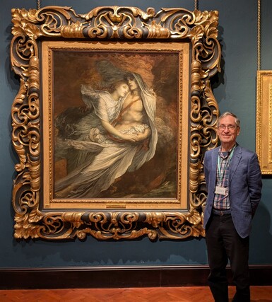 A wearing glasses and a blue jacket is standing next to a large painting by George Watts of Paolo and Francesca