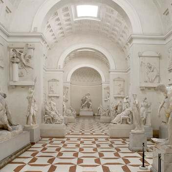 A collection of white classical sculptures are laid out round the edges of a large room with an arched round roof with white walls. The floor has brown and white tiling.