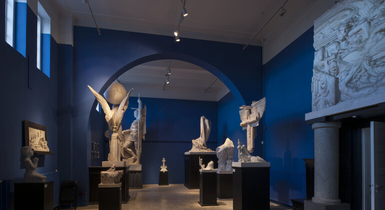 A collection of sculptures are laid out intermittently in a large room painted a dark blue